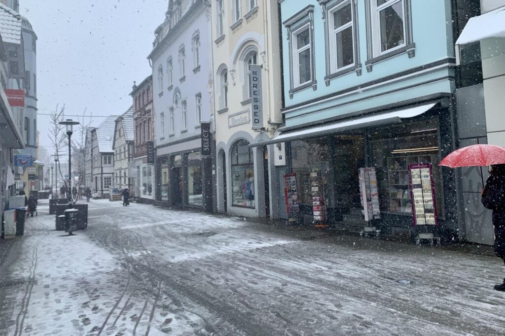 Zu sehen sind Läden in der Schwerter Innenstadt und eine Frau mit Regenschirm.