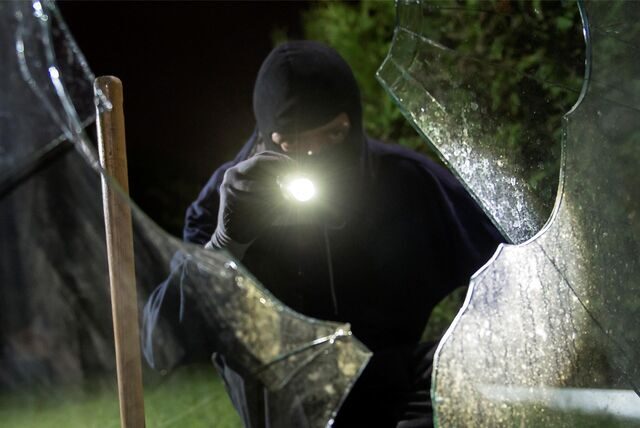 Einbrecher steht mit einer Taschenlampe vor einem eingeschlagenen Fenster