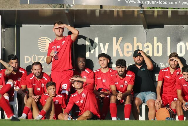 Spieler von Türkspor auf der Ersatzbank: Die Sonne scheint, mehrere halten sich eine Hand vor das Gesicht.