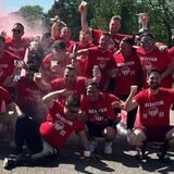 Die gesamte Fußballmannschaft sitzt jubelnd auf dem Boden. Sie heben die Hände in die Luft und feiern bei Sonnenstrahlen.