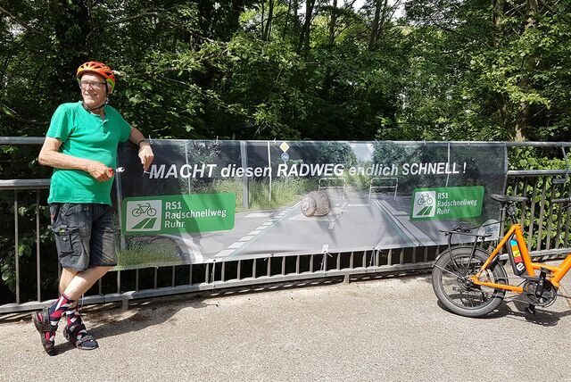 Hermann Strahl vom ADFC in Unna lehnt sich an ein Brückengeländer, an dem ein Protestbanner angebracht wurde. Es fordert eine schnellere Verwirklichung der Pläne für einen Radschnellweg durchs Ruhrgebiet.