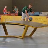 Zwei junge Männer spielen auf einer gewölbten Tischtennisplatte mit einem Handball. Der hintere Junge trägt ein grünes Trikot und der vordere ein blaues Trikot mit der Nummer 4.