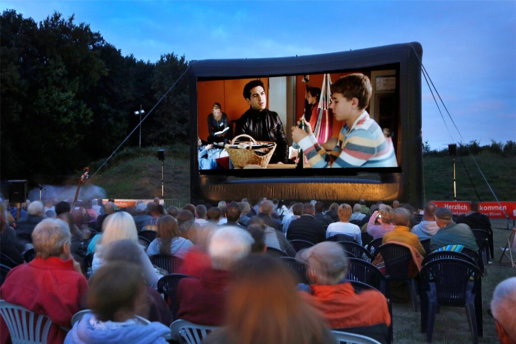 Menschen sitzen vor einer großen Leinwand im Freien und schauen einen Film. Menschen sitzen vor einer großen Leinwand im Freien und schauen einen Film.