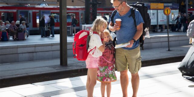 Ein Vater steht mit seinen zwei Töchtern am Bahnhof und zeigt ihnen einen Zettel.