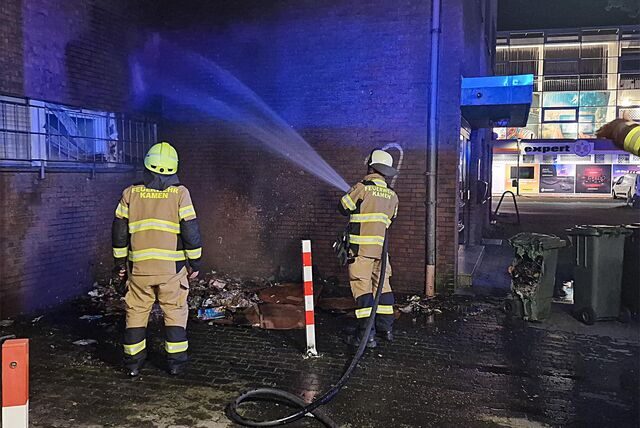 Zwei Feuerwehrmänner löschen in der Nacht eine Mülltonne, die an einer Hausfassade gebrannt hat. Auch zwei Fenster und Jalousien wurden beschädigt.