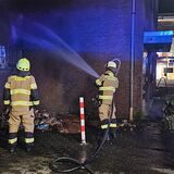 Zwei Feuerwehrmänner löschen in der Nacht eine Mülltonne, die an einer Hausfassade gebrannt hat. Auch zwei Fenster und Jalousien wurden beschädigt.