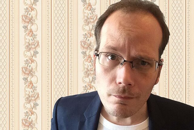 Foto von Comedian Mario Siegesmund vor einem gelben Hintergrund. Er trägt eine Brille und ein kariertes Hemd. Er spricht in ein Mikrofon und gestikuliert mit der linken Hand.