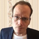 Foto von Comedian Mario Siegesmund vor einem gelben Hintergrund. Er trägt eine Brille und ein kariertes Hemd. Er spricht in ein Mikrofon und gestikuliert mit der linken Hand.