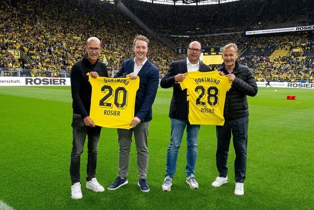 Vier Männer stehen auf dem Rasen eines vollen Stadions und präsentieren zwei Trikots.