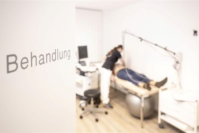 Ein Behandlungsraum beim Arzt. An der Tür steht das Wort „Behandlung“, im Hintergrund liegt ein Patient auf einer Liege, eine Ärztin steht davor.