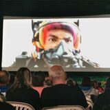 Eine Kinoleinwand, davor Publikum. Auf der Leinwand ein Moment des Films „Maverick“, während die Hauptfigur im Jet fliegt.