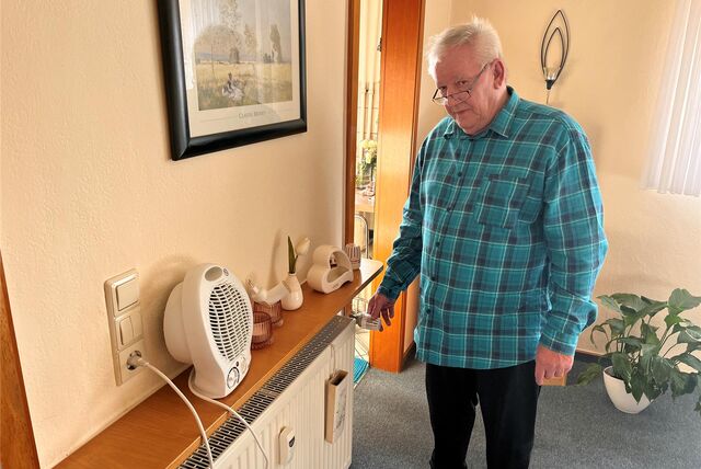 Mieter Helmut Bräckelmann steht im Oktober 2023 in seinem Wohnzimmer. Er hat eine Hand am Thermostat der ausgefallenen Heizung. Auf dem Sims darüber steht ein Heizlüfter.