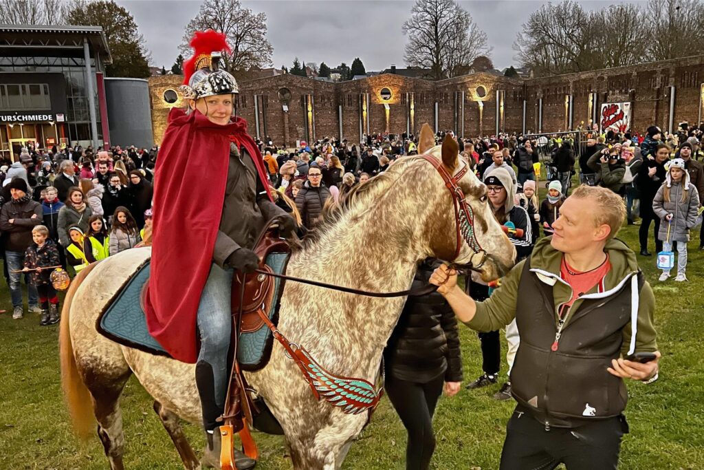 Eine Reiterin als Sankt Martin verkleidet sitzt auf einem Pferd. Im Hintergrund sind viele Kinder mit Laternen. Eine Reiterin als Sankt Martin verkleidet sitzt auf einem Pferd. Im Hintergrund sind viele Kinder mit Laternen.