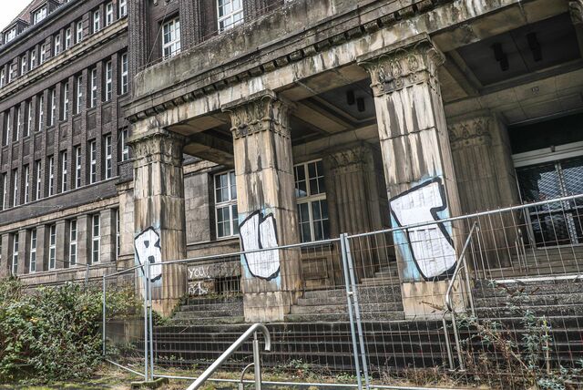 Der Eingang der ehemalige Hoesch-Zentrale in Dortmund von außen: ein Graffiti ist an Säulen vor dem Eingang gesprüht, ein Bauzaun steht vor der Treppe.