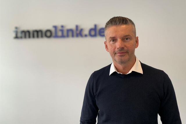 Der Makler Anel Kicin steht in seinem Büro. Im Hintergrund ist eine Schriftzug mit dem Namen seines Unternehmens zu sehen: Immolink.de