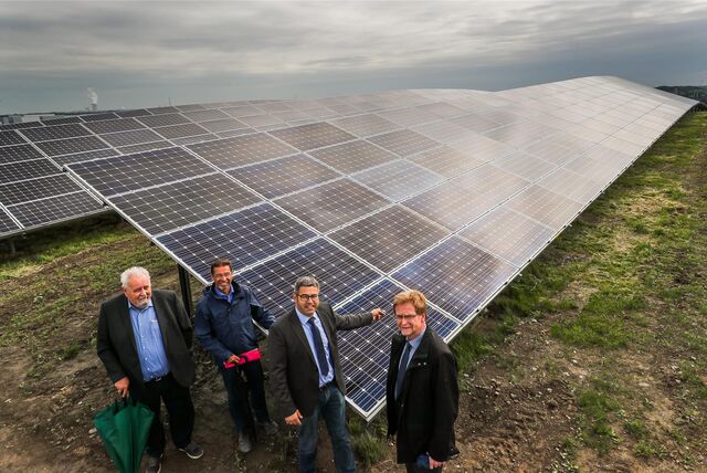 Vier Männer stehen vor riesigen Solarpaneelen auf einer Halde. Im Hintergrund ist ein Kraftwerk zu sehen.
