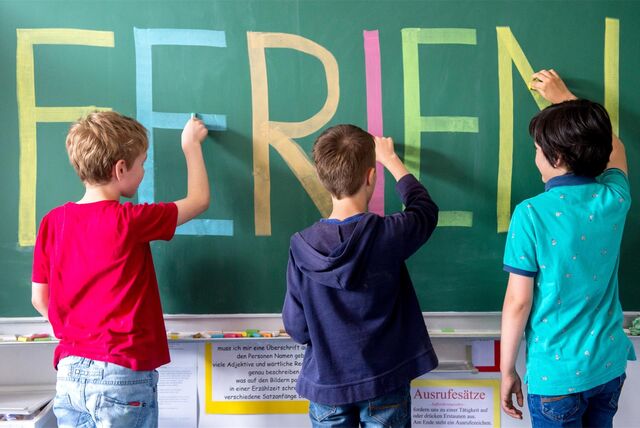 Schulkinder schreiben mit bunter Kreide das Wort „Ferien“ an eine Tafel.