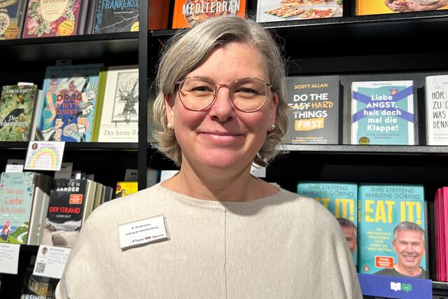 Die Filialleiterin einer Buchhandlung steht vor einem Regal mit vielen bunten Büchern. Sie hat kurze, hellblonde Haare, trägt eine Brille und einen beigefarbenen Pullover. Sie lächelt in die Kamera.