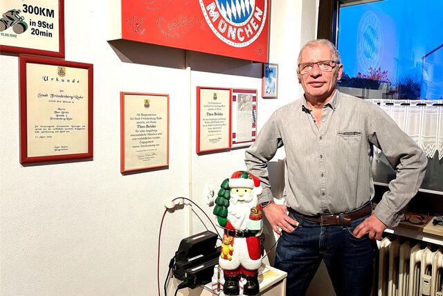 Ein Mann steht hinter einer Weihnachtsmann-Figur neben einer Wand mit Urkunden und Bayern München-Logo.