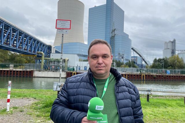 Ein Mann steht mit einem Mikrofon in der Hand am Ufer des Kanals in Datteln, im Hintergrund ein Kraftwerk.