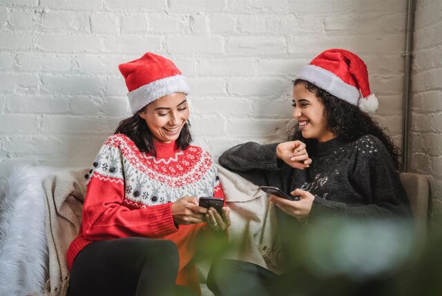 Zwei Frauen mit Nikolausmützen sitzen lachend auf einem Sofa und schauen auf ihre Smartphones.