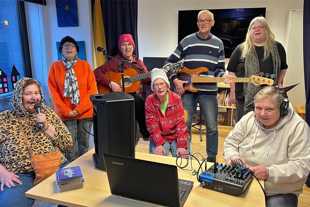 Eine Gruppe der Caritas-Kontaktstelle sitzt an einem Tisch, auf dem ein Laptop, eine Soundbox und ein Mischpult stehen. Die Menschen halten Mikros und Instrumente in den Händen.