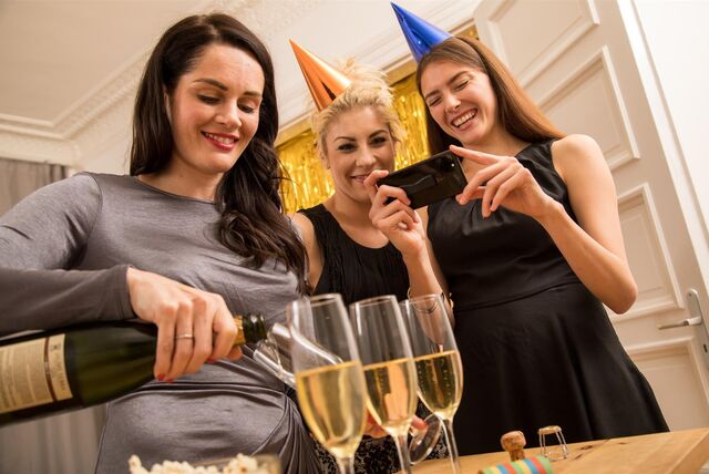 Drei Frauen auf einer Silvesterparty. Eine Frau gießt Sekt in Gläser ein, eine andere hält ein Smartphone in der Hand.