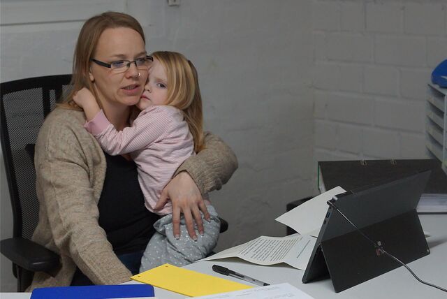 Manuela Thiele sitzt mit ihrer Tochter auf dem Arm auf einem Schreibtischstuhl, vor sich auf dem Tisch Unterlagen und ein aufgestelltes Tablet.
