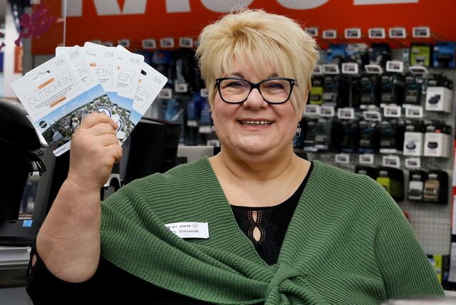 Eine Frau mit kurzen blonden Haaren und schwarzer Brille schaut in die Kamera und hält einen Fächer aus Gutscheinen in die Höhe. Sie trägt ein schwarzes Oberteil und darüber einen jägergrünen Strick-Poncho.