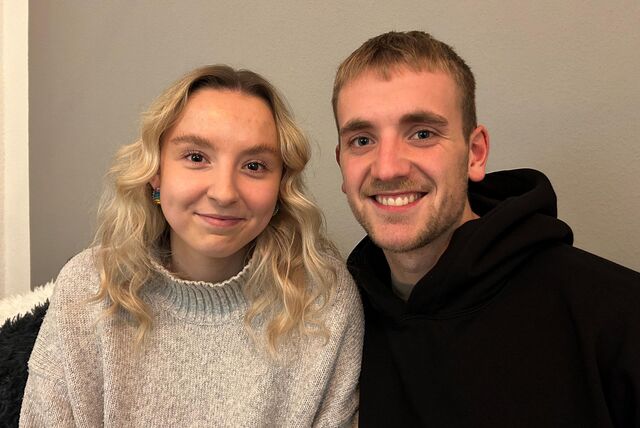 Ein junges Paar schaut in die Kamera und lächelt. Sie (links im Bild) hat mittellanges hellblonde Haar, braune Augen und trägt einen beigefarbenen Pullover. Er hat kurzes, mittelblondes Haar, blaue Augen und trägt einen schwarzen Kapuzenpulli.