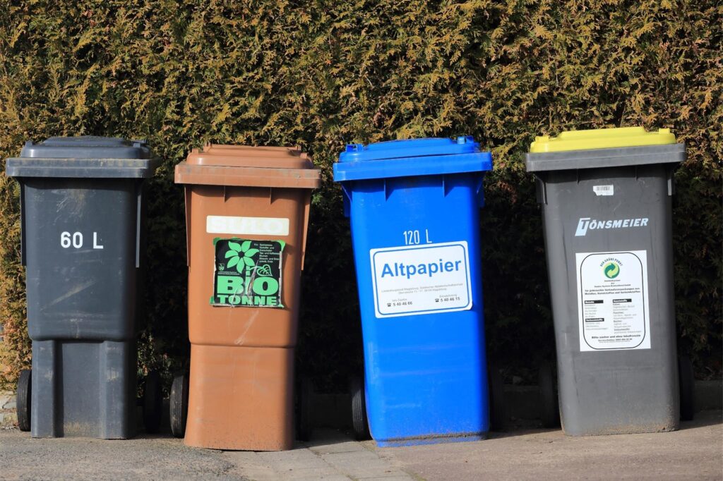 Mülltonnen für Hausmüll (schwarz), Bioabfälle (braun), Altpapier (blau) und Wertstoffe (gelb) stehen vor einer Hecke. Mülltonnen für Hausmüll (schwarz), Bioabfälle (braun), Altpapier (blau) und Wertstoffe (gelb) stehen vor einer Hecke.