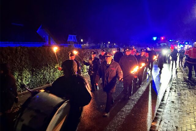 Im Regen sieht man einen langen Zug mit Musikern, Feuerwehrleuten mit Fackeln und im Hintergrund Autos.
