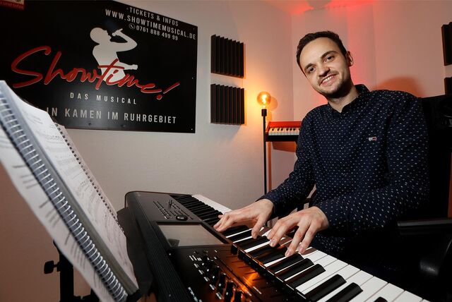 Ein Musiker sitzt an einem Keyboard. Die Finger auf den Tasten, den Blick in Richtung Kamera. Vor ihm ein Notenständer mit Noten. Im Hintergrund ein Poster des Musicals: Showtime.