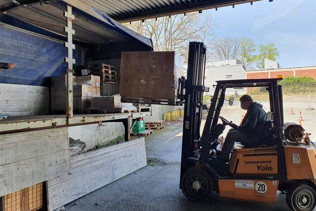Ein Mann sitzt in einem orangen Gabelstapler und lädt eine Palette mit Paketen in einen seitlich geöffneten Lkw-Anhänger.