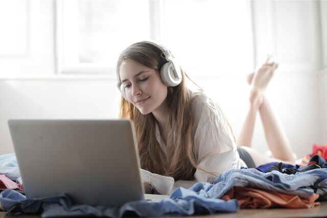 Eine junge Frau liegt mit ihrem Laptop auf ihrem Bett und hat dabei Kopfhörer auf. Sie hört augenscheinlich etwas über die Kopfhörer.