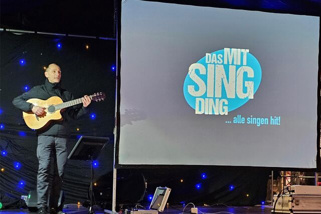 Stefan Nussbaum steht mit seiner Gitarre auf der Bühne, auf der Leinwand im Hintergrund ist.