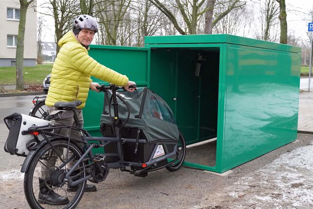 Ein Mann mit gelber Jacke und Helm auf dem Kopf schiebt ein Lastenrad in eine der Lastenradboxen.