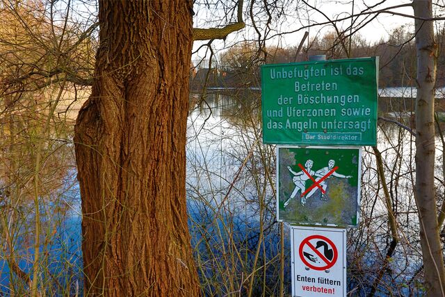 Drei Warnschilder an einem See weisen darauf hin, dass das Schlittschuhlaufen, Entenfüttern, Betretung der Böschung und das Angeln verboten sind.