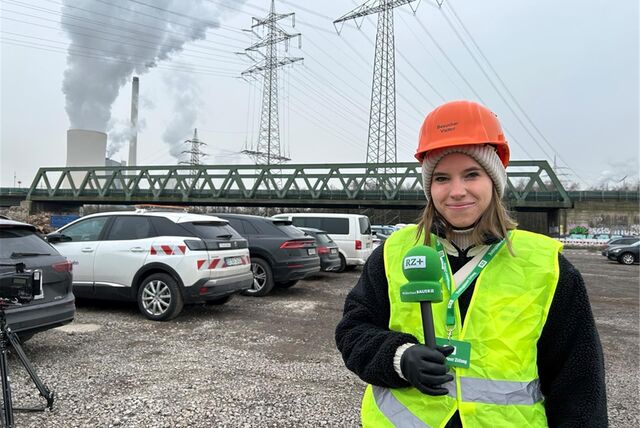Reporterin Janine Jähnichen mit Helm und Mikro auf einer Baustelle. Im Hintergrund sind eine Brücke und ein Kraftwerk zu sehen.