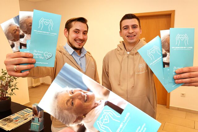Zwei junge Männer, die mehrere Flyer in Richtung Kamera halten. Sie lächeln und tragen beigefarbene Stoffjacken, auf denen das Symbol ihres Unternehmens zu sehen ist: Ein Herz mit Herzschlag, das von zwei Händen gehalten wird.