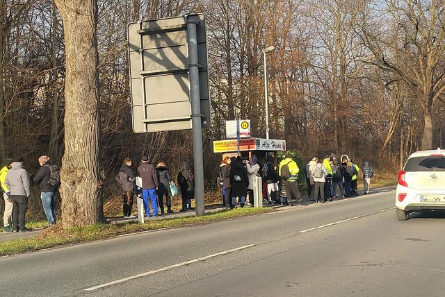 An der Bushaltestelle Alte Heide in Unna stehen zahlreiche Menschen, die auf den Bus warten. Ein weißes Auto fährt vorbei