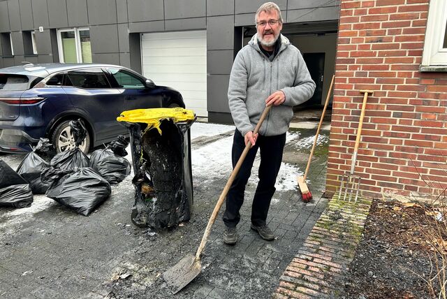 Ein älterer Mann steht in einer Garageneinfahrt. Er hat einen Spaten in der Hand, neben ihm eine vom Feuer zerstörte Mülltonne. Er schaut in die Kamera, im Hintergrund sieht man mehrere schwarze Müllsäcke.