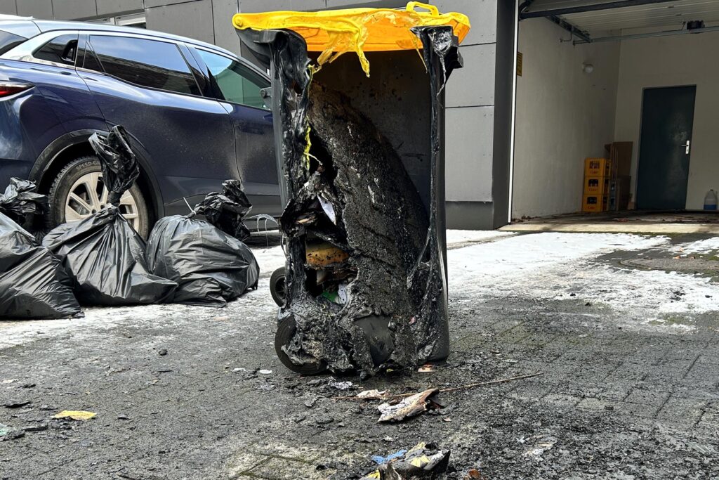 Eine gelbe Tonne in einer Einfahrt. Im Hintergrund liegen Müllsäcke und es steht ein Auto dort. Die Mülltonne ist durch das Feuer geschmolzen. Eine gelbe Tonne in einer Einfahrt. Im Hintergrund liegen Müllsäcke und es steht ein Auto dort. Die Mülltonne ist durch das Feuer geschmolzen.