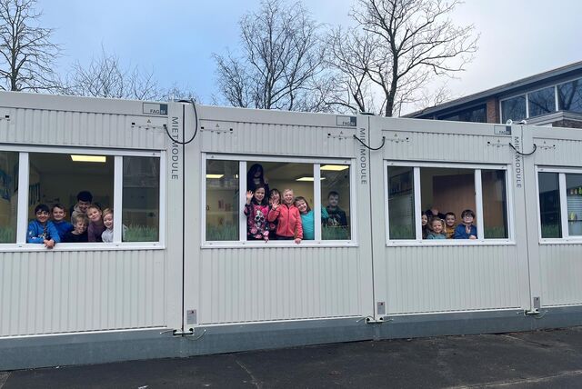 Kinder winken aus dem Fenster eines Klassenzimmers, das sich schon jetzt in einem Container befindet.