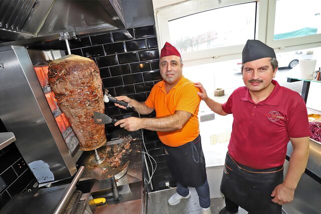 Zwei Männer stehen in einem Imbiss am Dönerspieß. Der eine schneidet gerade Fleisch ab, der andere hält seine rechte Hand auf die Schulter seines Kollegen. Beide schauen in die Kamera.