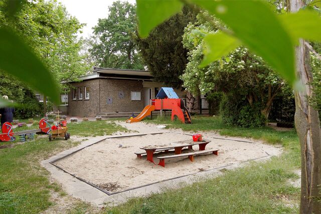 Vorn im Bild ist ein Sandkasten zu sehen, dahinter steht die rote Rutsche und dahinter das flache Gebäude der Kita Flohzirkus.