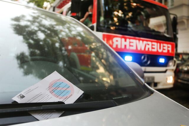 Ein „Knöllchen“ klemmt hinter dem Scheibenwischer eines Autos. Im Hintergrund ist ein Feuerwehrfahrzeug mit eingeschaltetem Blaulicht