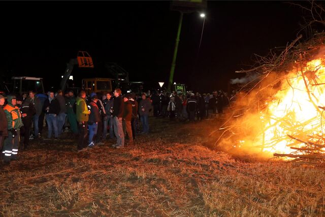 Rechts brennt ein Haufen Holz. Auf der linken Seite stehen Menschen und reden. Im Hintergrund sind Trecker zu sehen.