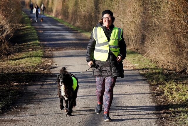 Eine Frau sitzt in der Hocke auf einem Weg. Neben ihr sitzt ein schwarz-weißer Hund. Hund und Mensch tragen eine gelbe Warnweste.