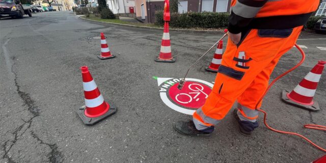 Ein Mann in oranger Warnkleidung hält einen Flammenwerfer an ein Piktogramm auf einer Straße, auf dem „Radring“ steht. Rot-weiße Pylonen umgeben die Baustelle
